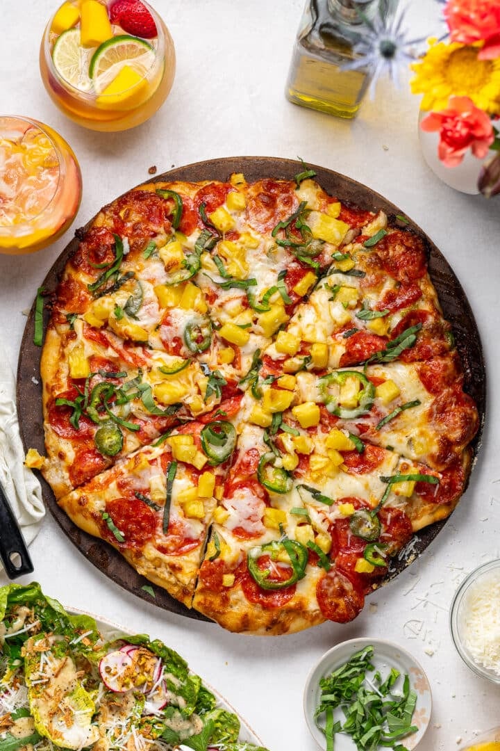 Sliced pepperoni pineapple jalapeño pizza with melted mozzarella and fresh basil, served with salad and glasses of sangria