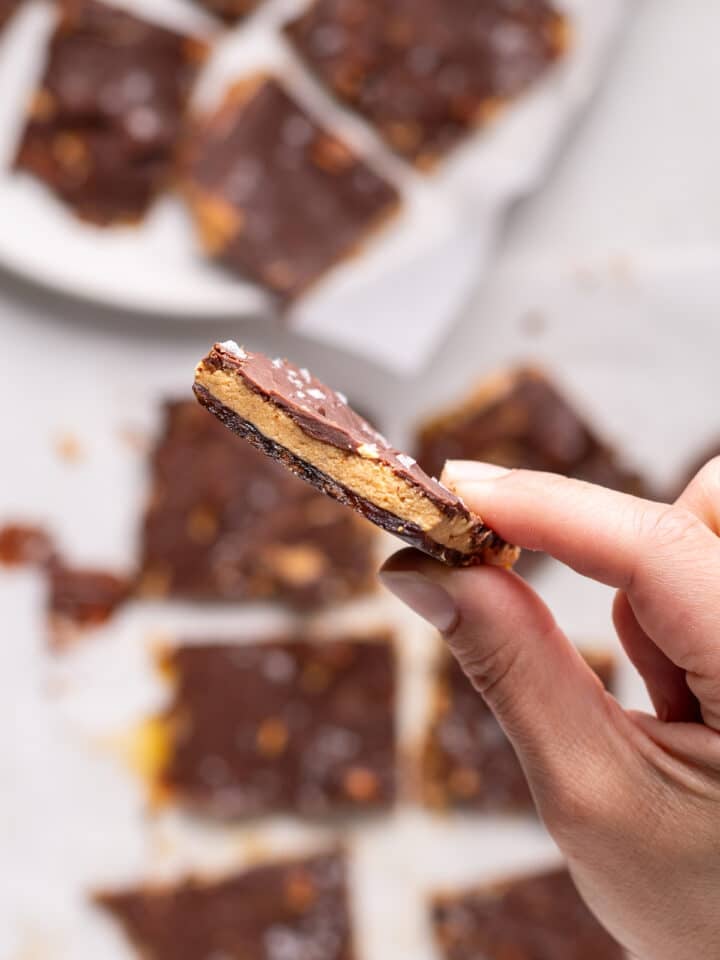A hand holding a thin piece of peanut butter date bark with chocolate, peanuts, and flaky sea salt