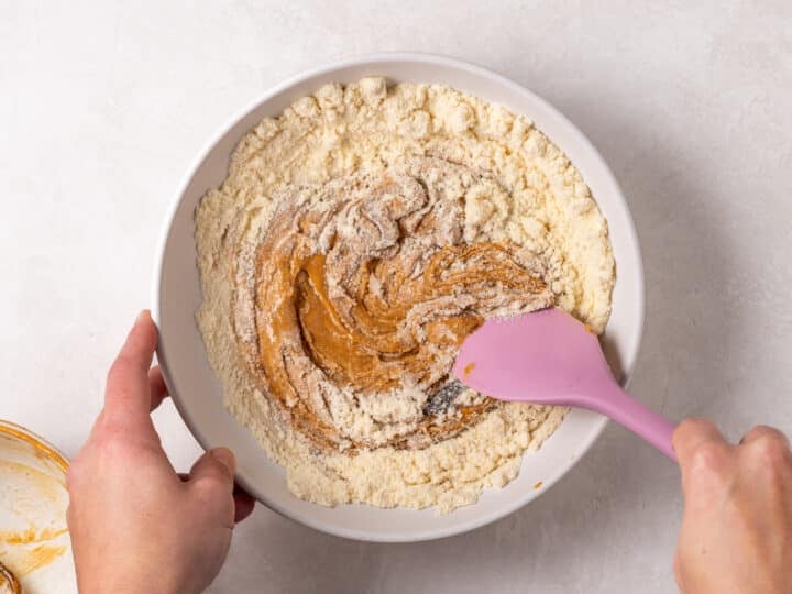 A pink spatula stirring peanut butter, protein powder, vanilla bean paste, and pure maple syrup together in a mixing bowl