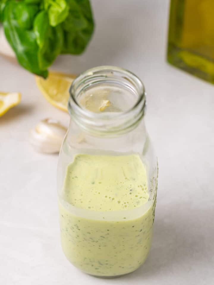 Glass jar filled with creamy pesto salad dressing beside fresh basil, lemon, garlic, and olive oil.