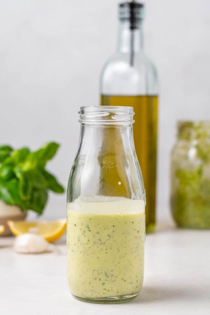 Creamy pesto vinaigrette in a glass jar with basil, lemon, garlic, olive oil, and pesto in the background.