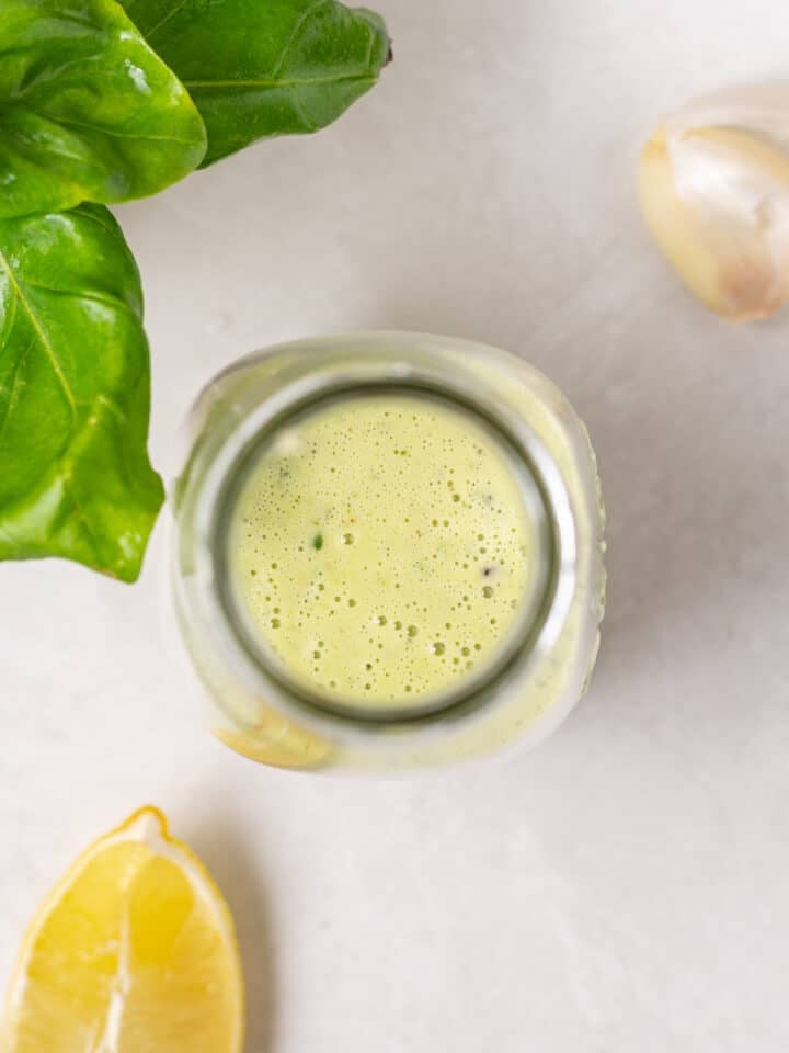 Overhead view of pesto vinaigrette dressing in a glass jar with fresh basil, lemon, and garlic on a light countertop.