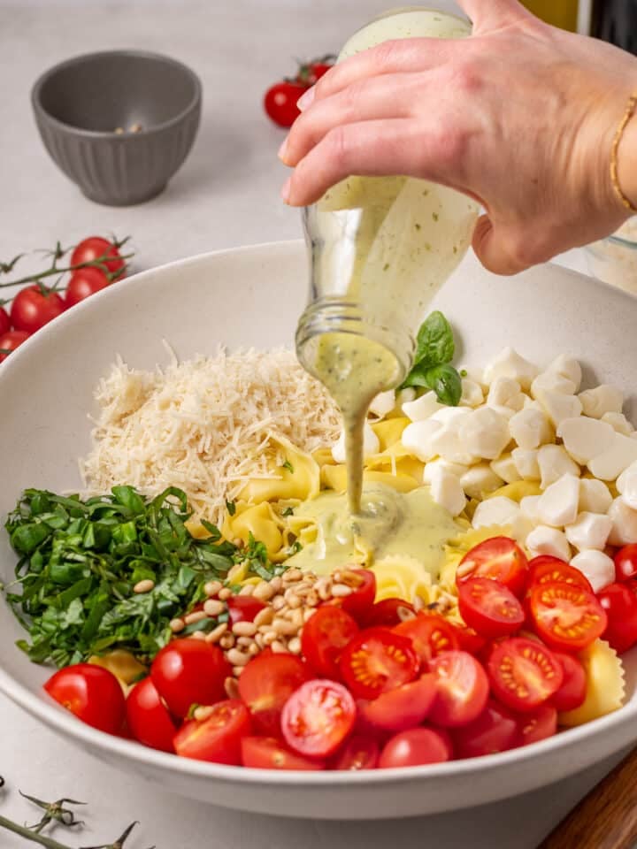 Creamy pesto vinaigrette being poured over tortellini salad with cherry tomatoes, mozzarella, basil, pine nuts, and parmesan.
