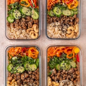 Four meal prep containers filled with rice, saucy chicken, cucumbers, carrots, peppers, cashews and sesame seeds.