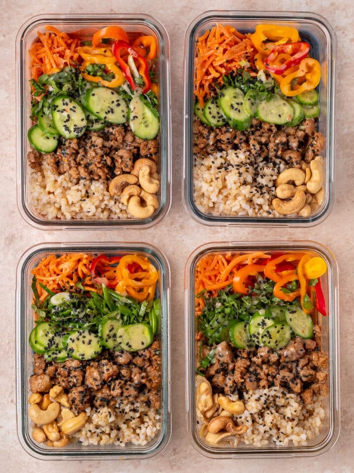 Four meal prep containers filled with rice, saucy chicken, cucumbers, carrots, peppers, cashews and sesame seeds.