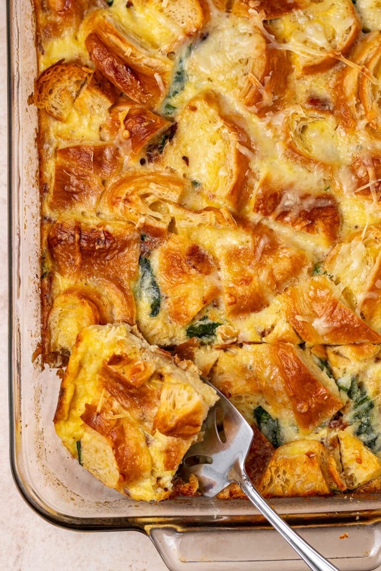 Slice of high protein egg bake with bacon, leafy greens, and croissants being lifted from a baking dish with a metal spatula.