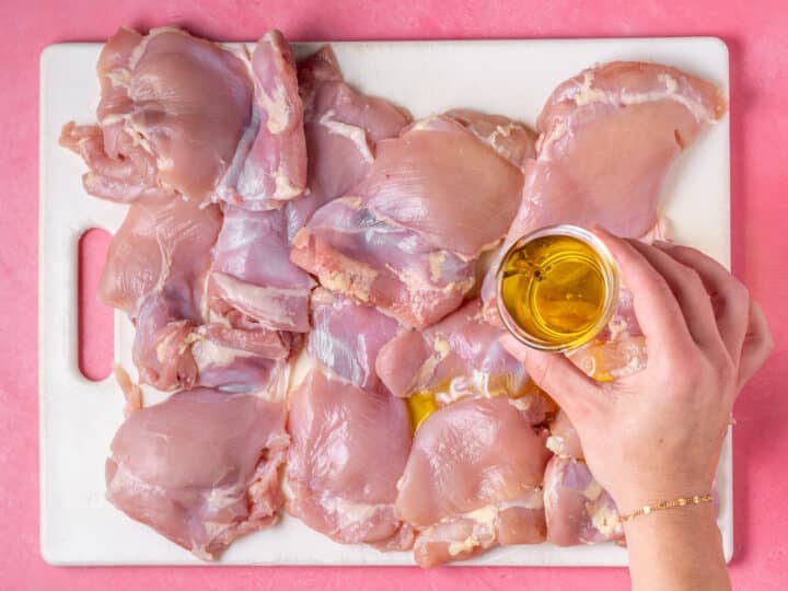 Olive oil being drizzled over raw chicken thighs on a cutting board.