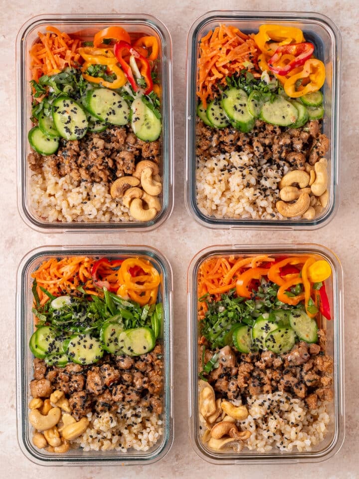 Four meal prep power bowls with rice, chicken, cucumbers, carrots, peppers, cashews, herbs and sesame seeds.
