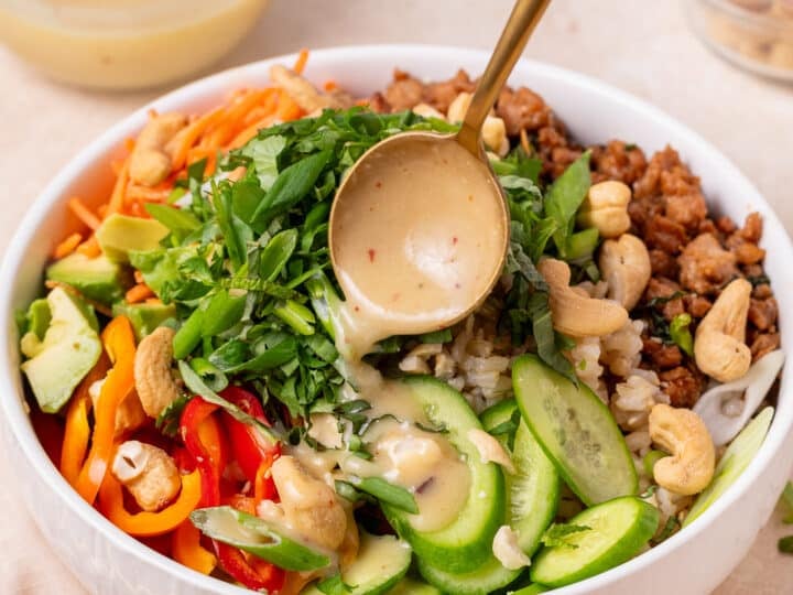 Creamy miso vinaigrette being drizzled over a rice bowl with garlic ginger chicken, cucumbers, peppers, herbs and cashews.