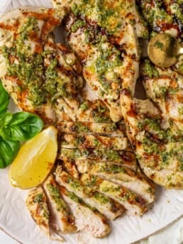 Sliced pesto chicken breast with basil pesto, balsamic glaze, fresh basil, and lemon on a serving platter.