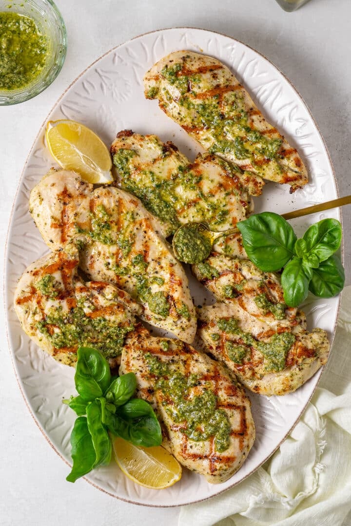 large white platter with grilled chicken dolloped with basil sauce, lemon wedges and fresh basil