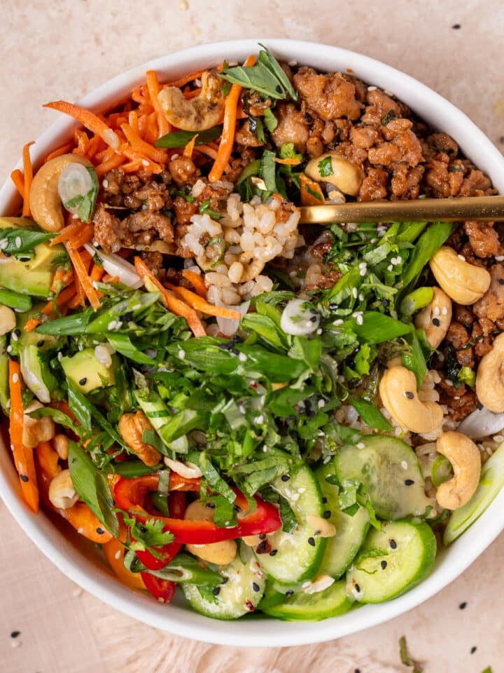 Close-up of a power bowl with garlic ginger chicken, brown rice, avocado, fresh herbs, cashews, cucumbers and a forkful of rice.