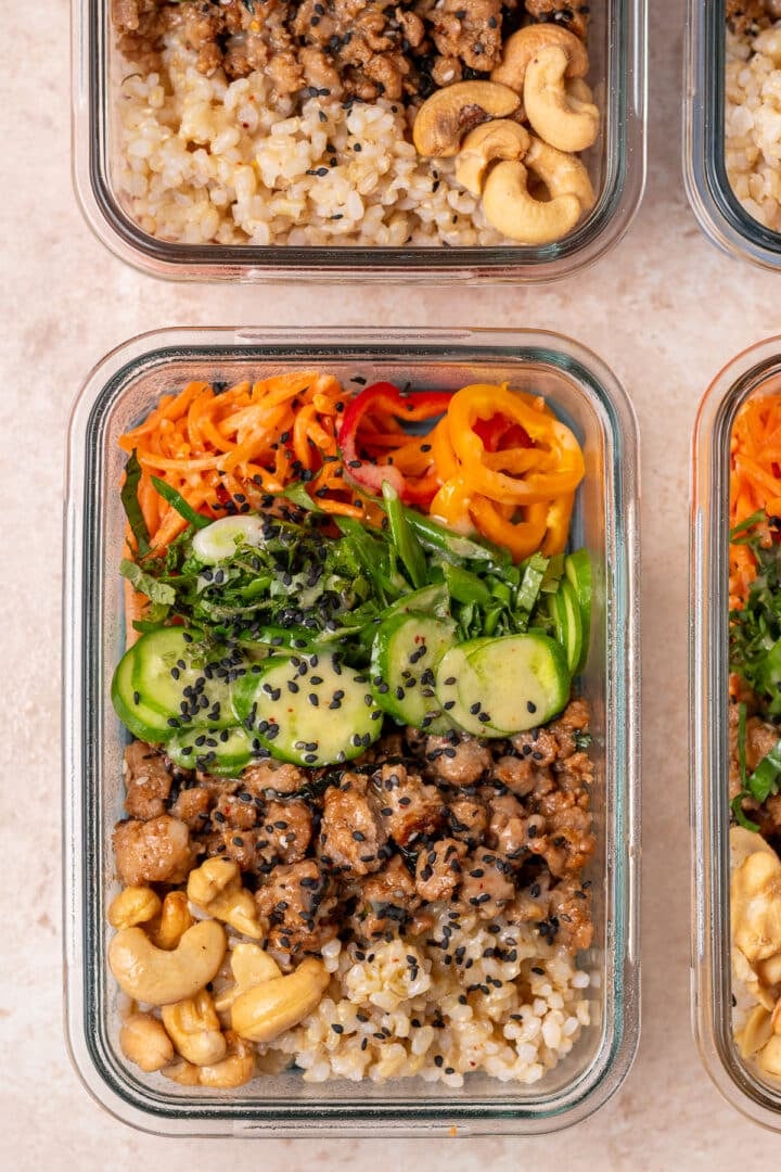Close-up of a meal prep container with brown rice, chicken, cucumbers, carrots, peppers, cashews and black sesame seeds.
