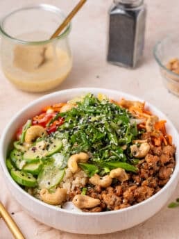 Chicken power bowl with brown rice, cucumbers, carrots, peppers, herbs, cashews and miso vinaigrette in the background.