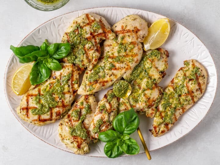 Grilled chicken with pesto, fresh basil, and lemon wedges on a white serving platter