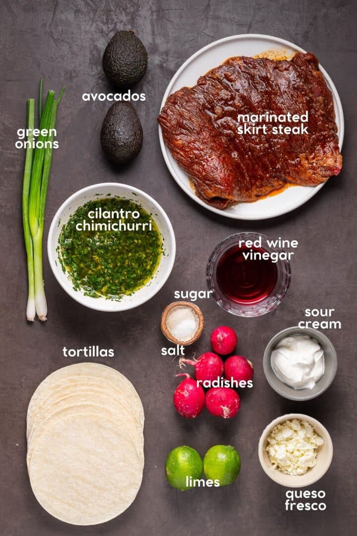 Ingredients for recipe including marinated steak, tortillas, avocado, radishes, limes, chimichurri, sour cream, queso fresco and green onions.
