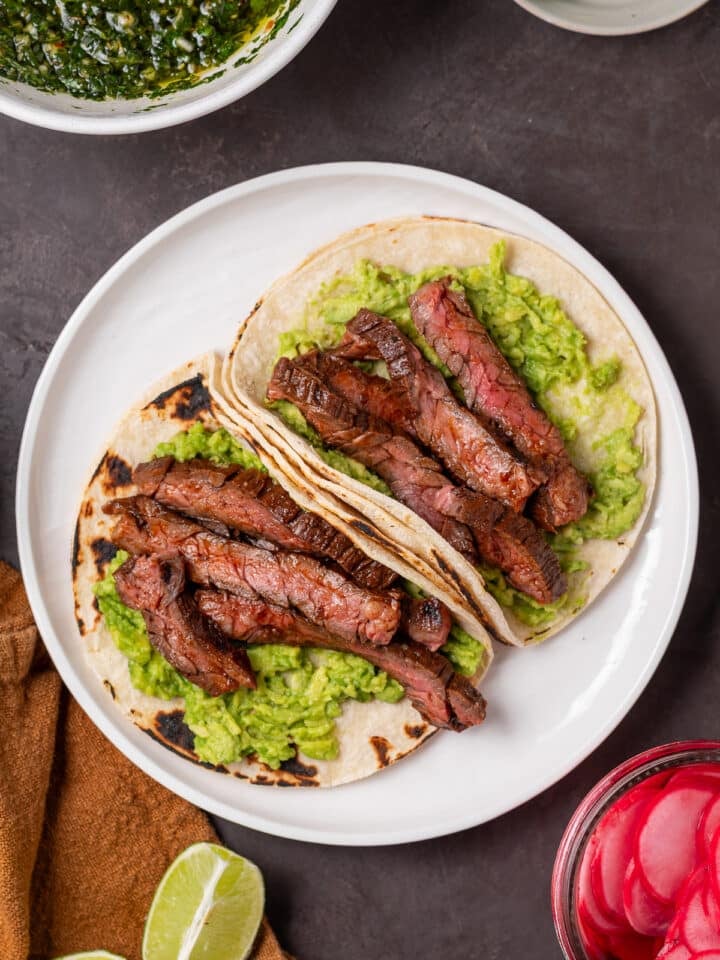 Overhead view of marinated skirt steak layered over mashed avocado in warm tortillas.