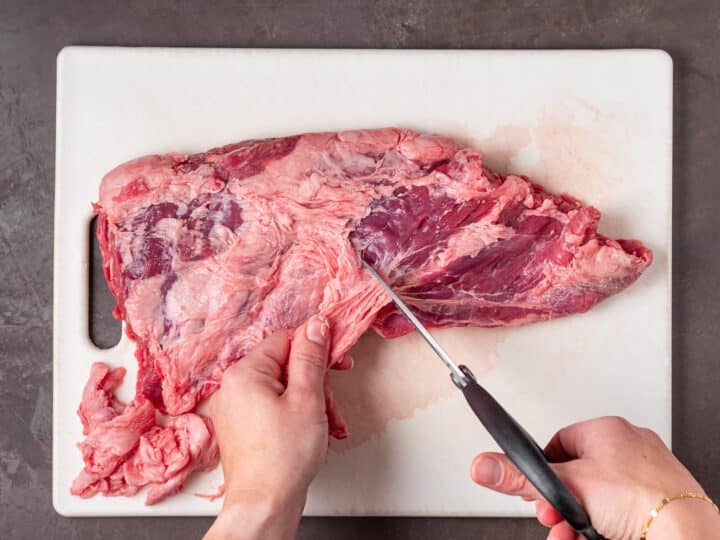 Raw steak being trimmed with kitchen scissors on a white cutting board.