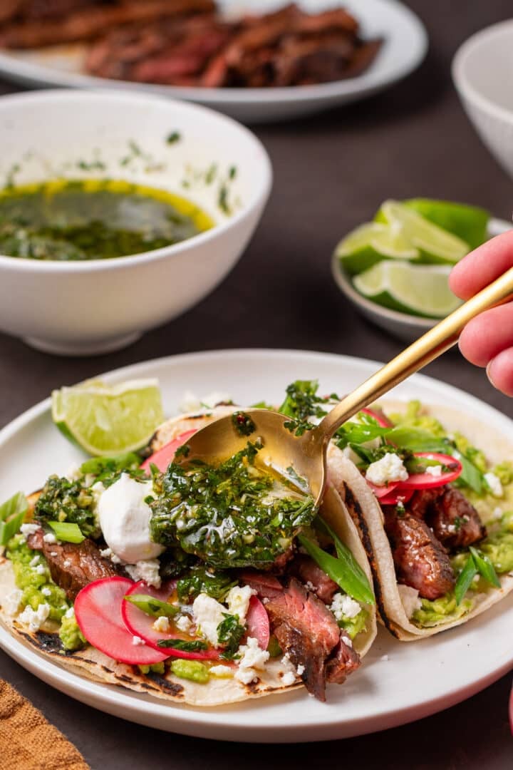 Cilantro chimichurri being spooned over skirt steak tacos with avocado, pickled radishes, queso fresco and sour cream.