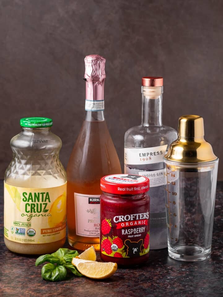 A shaker and ingredients for an easy cocktail including gin, prosecco, raspberry jam, lemon juice and fresh basil.