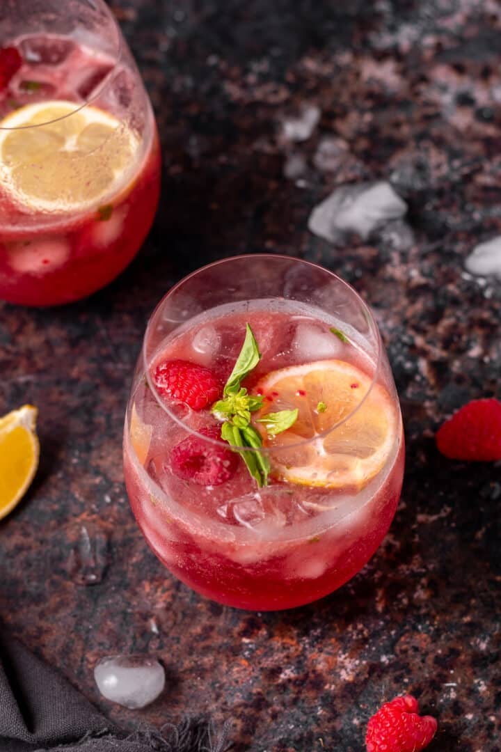 Two pink gin spritz cocktails over ice garnished with raspberries, lemon slices and fresh basil.