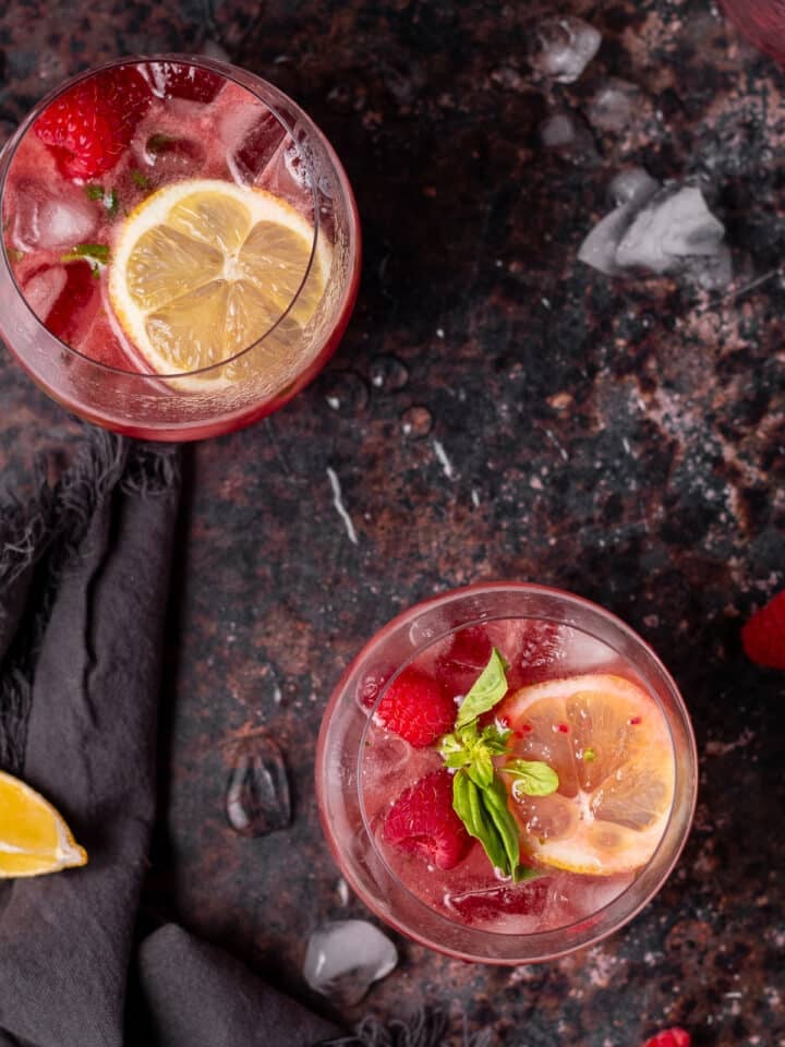 Overhead view of two fruity gin spritz cocktails with raspberries, basil and lemon slices.