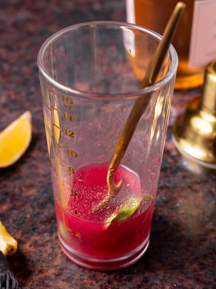 Basil leaves, raspberry jam, lemon juice and gin being muddled together with a spoon.