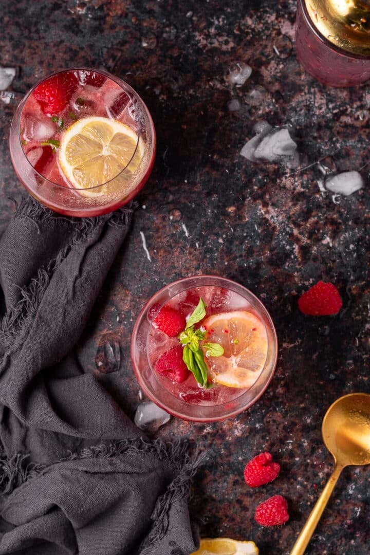 Overhead view of two cocktails garnished with lemon, basil and fresh raspberries.