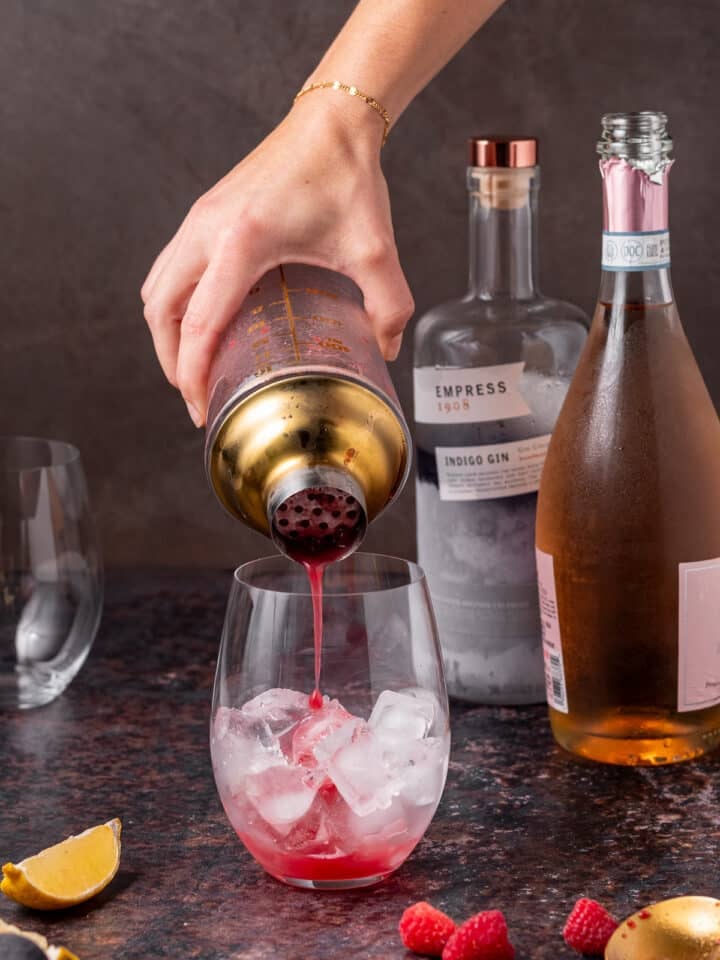 Raspberry gin mixture being strained from a cocktail shaker into a glass filled with ice.
