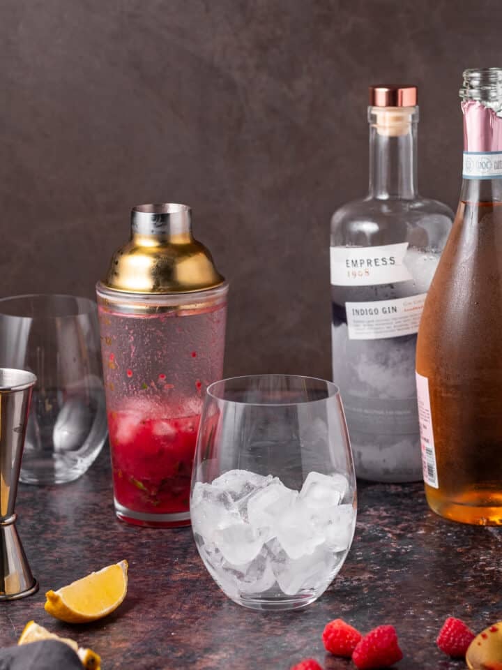 Cocktail shaker filled with raspberry jam, basil, lemon and gin beside a glass of ice.