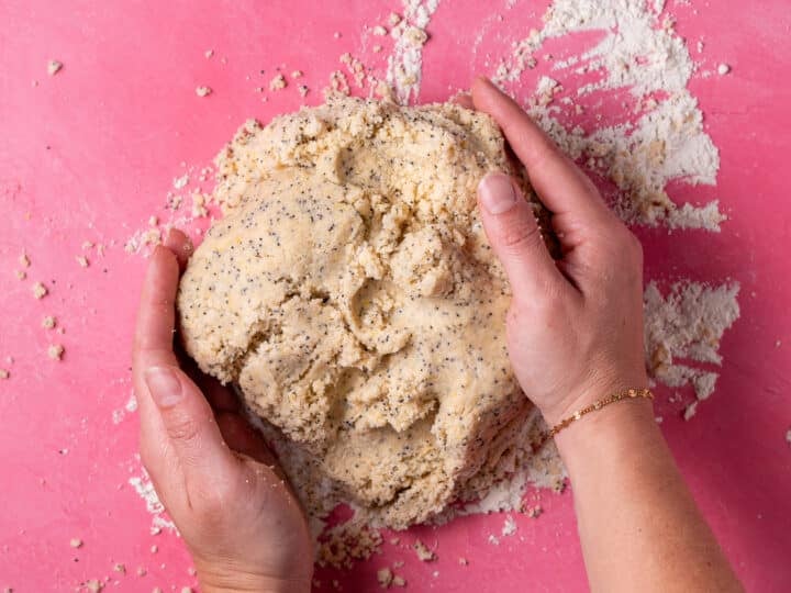 Hands gathering crumbly scone dough together on a lightly floured surface.