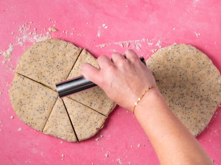 Lemon poppyseed scone dough being cut into wedges with a metal bench scraper.
