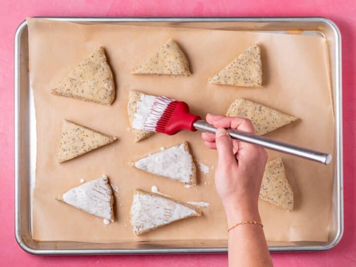 Unbaked lemon poppyseed scones being brushed with heavy cream on a parchment-lined baking sheet.
