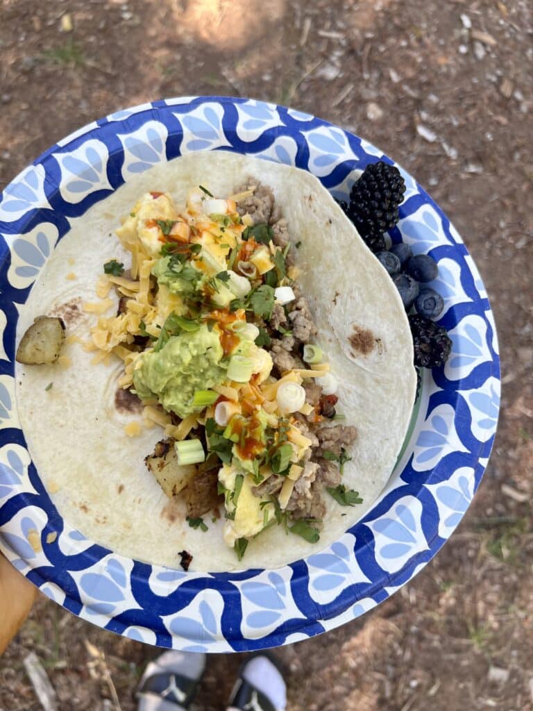 Breakfast burrito filled with eggs, potatoes, sausage, cheddar, avocado, green onions, cilantro and hot sauce on a paper plate at the campsite