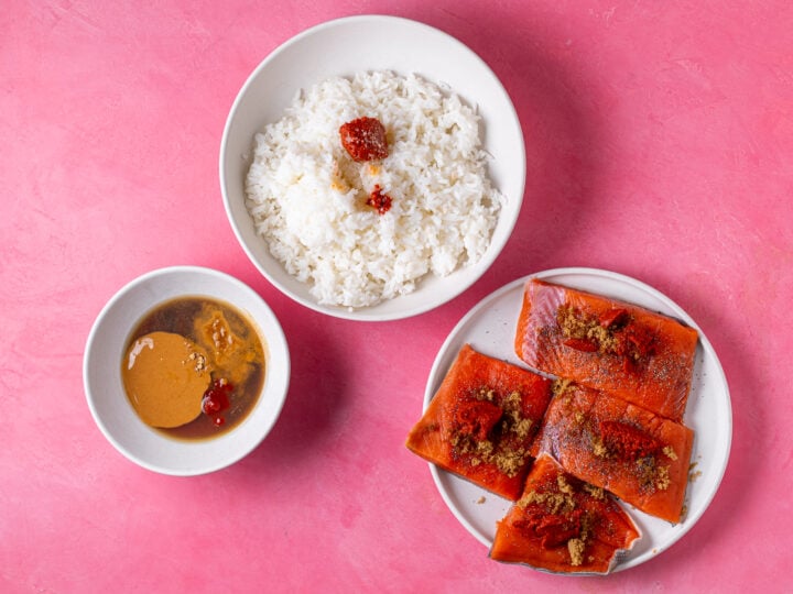 Cooked jasmine rice with red curry paste and peanut sauce ingredients in separate bowls before mixing and seasoned salmon fillets on a plate