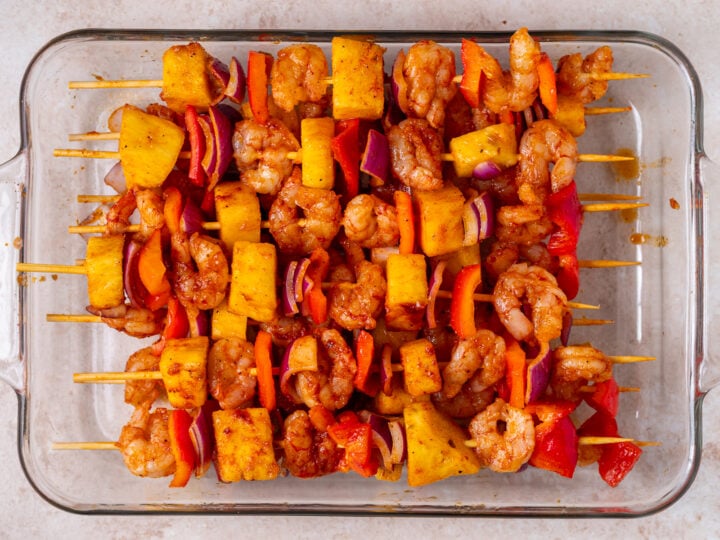 Assembled shrimp pineapple skewers with red bell pepper and red onion arranged in a glass baking dish.