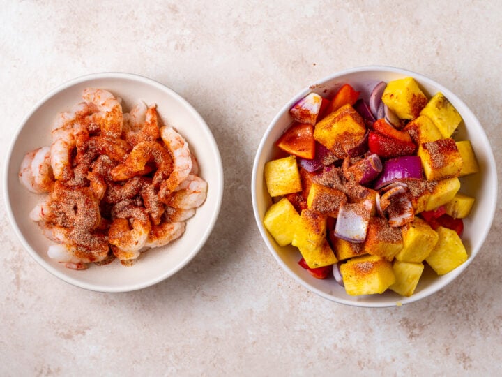 Shrimp in one bowl and chopped pineapple, red bell pepper and red onion in another bowl with seasoning blend on top.