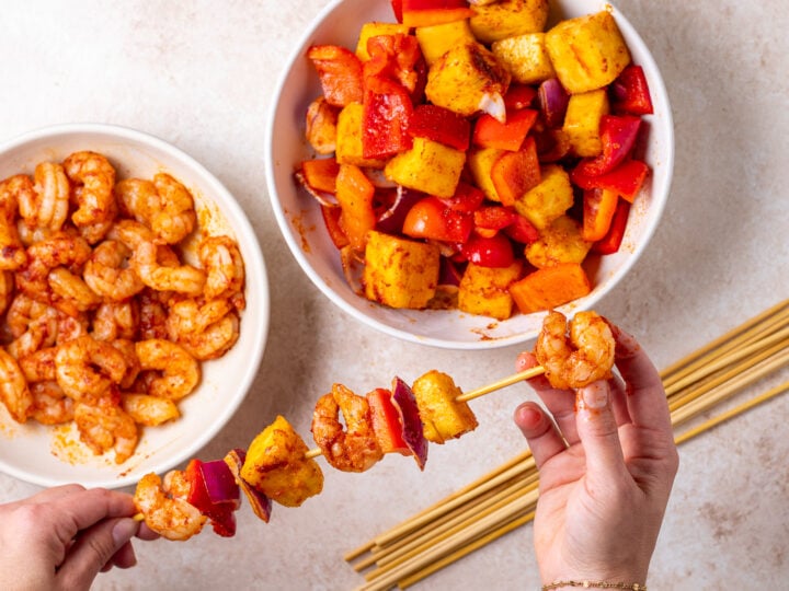 Hands threading seasoned shrimp, pineapple, red bell pepper and red onion onto a bamboo skewer.