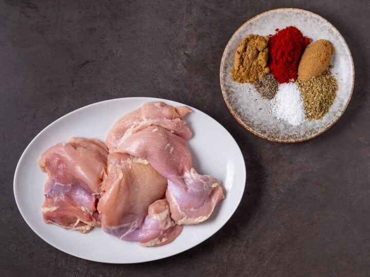 Raw chicken thighs beside a seasoning blend of cumin, smoked paprika, brown sugar, oregano, salt and black pepper.