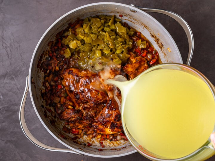 Diced green chiles and chicken broth being poured over seared chicken thighs and sautéed vegetables in a large pot.