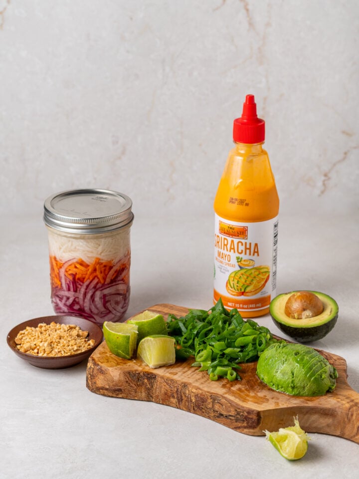 Ingredients for banh mi smash tacos toppings, including jar of quick-pickled veggies, avocado, green onions, crush peanuts, sriracha mayo, and lime wedges.