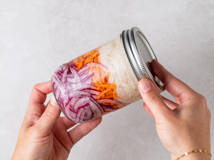 Glass mason jar filled with julienned daikon radish, shredded carrots and thinly sliced red onion for quick pickling.