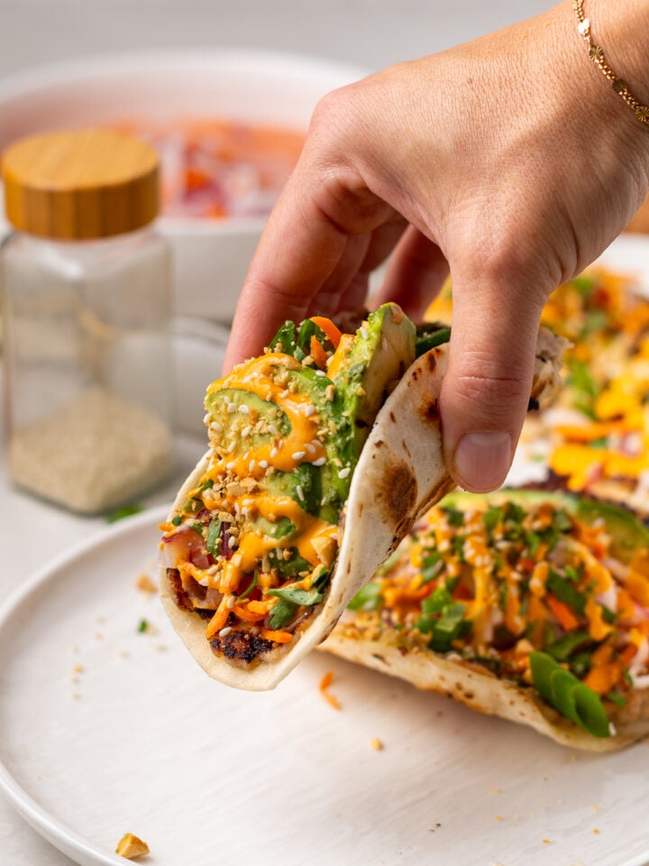 Hand holding a folded banh mi taco with sriracha mayo and sesame seeds on top.