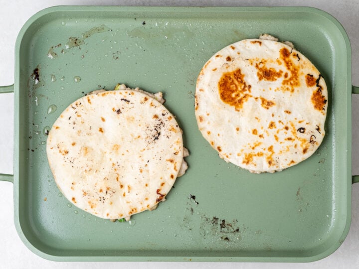 Two ground chicken smash tacos cooking tortilla-side up on a nonstick griddle.