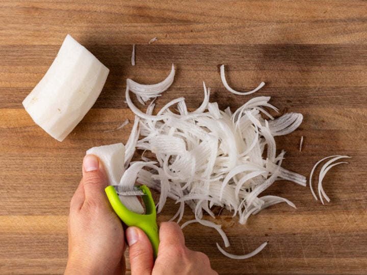 Using a julienne peeler to cut daikon radish into thin strips on a wooden cutting board.