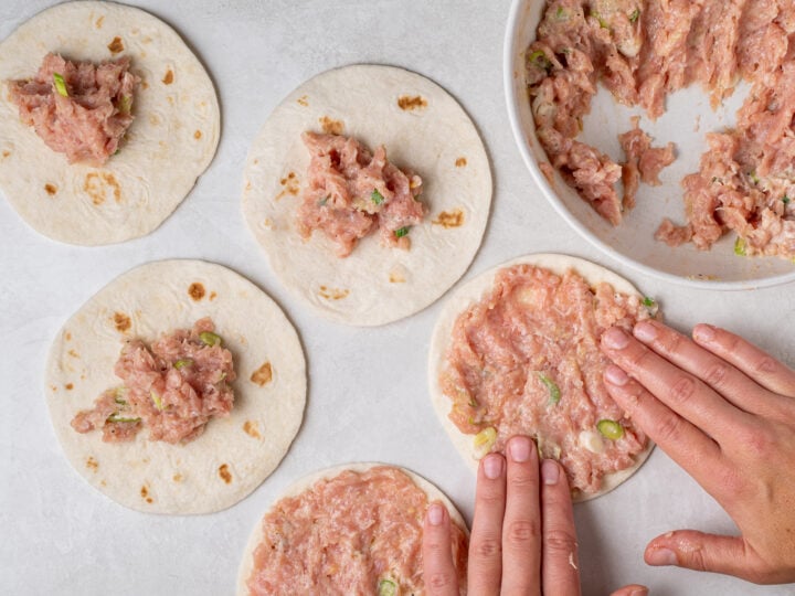 Hands spreading seasoned ground chicken in a thin layer across small flour tortillas.