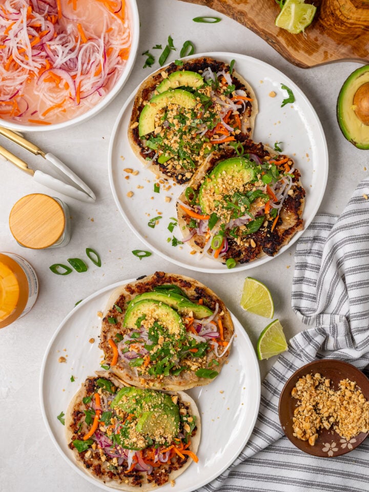 Four banh mi smash tacos on white plates with pickled vegetables, fresh herbs, avocado and crushed peanuts.