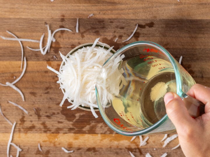Vinegar pickling mixture in liquid measuring cup being poured over shredded veggies in a glass jar.