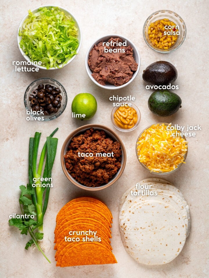 Overhead view of labeled ingredients for homemade gordita crunch tacos, including taco meat, refried beans, shredded cheese, tortillas, crunchy shells and toppings.