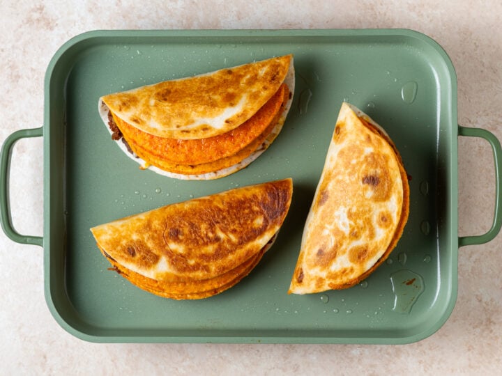 Three cheesy gordita crunch taco shells being toasted on a green griddle pan.